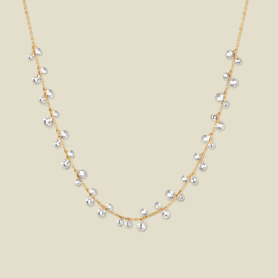 A dainty gold chain necklace with clusters of clear, round crystals dangling off, shown against a solid beige background.
