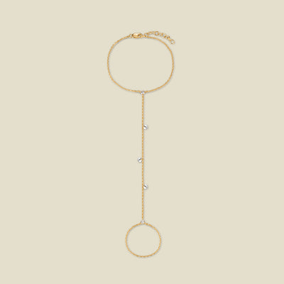 A dainty gold hand chain featuring small round clear crystals dangling throughout, shown against a solid beige background.