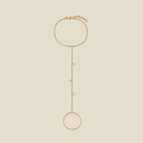 A dainty gold hand chain featuring small round clear crystals dangling throughout, shown against a solid beige background.