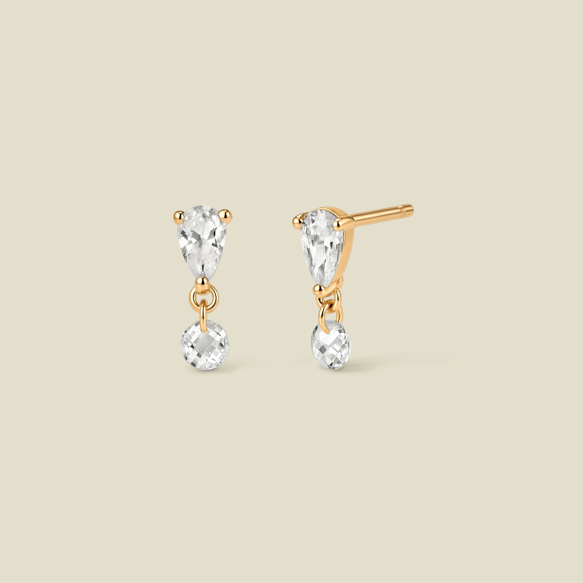 A pair of gold stud earrings featuring an upside down tear-drop shaped clear stone, and a small clear round crystal hanging from each, shown on a solid beige background.