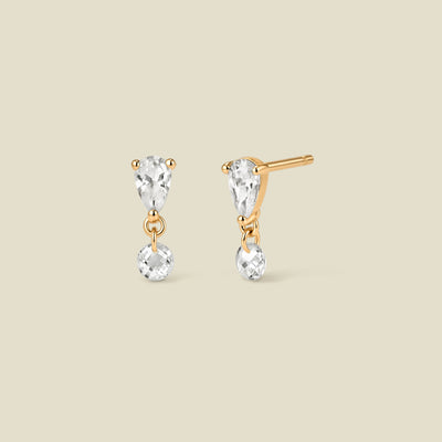 A pair of gold stud earrings featuring an upside down tear-drop shaped clear stone, and a small clear round crystal hanging from each, shown on a solid beige background.