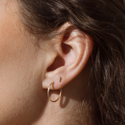 Close-up of an ear wearing a thin gold hoop earring and a small vertical bar stud with pave diamonds.