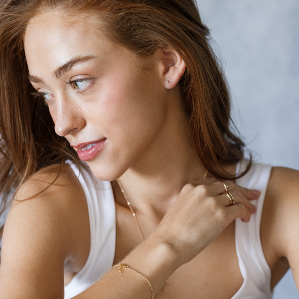 A lifestyle image of a woman wearing a white tank top and gold jewelry including a bracelet, necklace, rings, and a stud earring.
