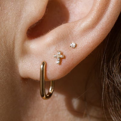 Close-up of an ear with gold hoop earring, cross-shaped stud, and small round diamond earring.