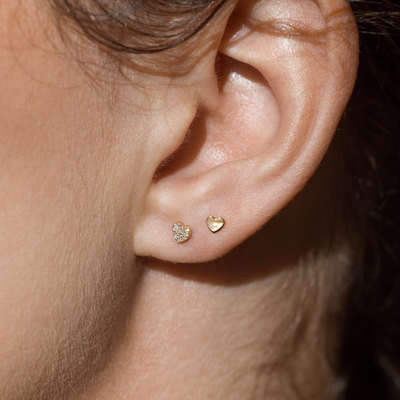 Close up of an ear wearing two heart shaped gold stud earrings, one with diamonds.