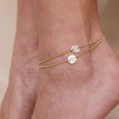 A close-up of a person's ankle wearing two gold chain anklets each with a circular disc, one engraved with a sun and the other with a wave design.