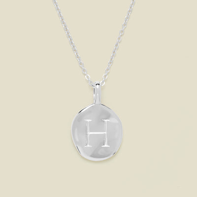 Silver necklace with a round pendant engraved with the letter 'H' on a beige background