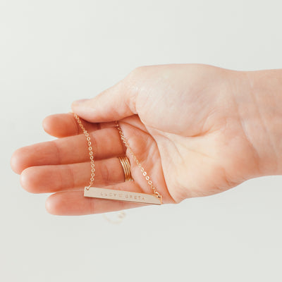 A hand holding a delicate gold horizontal bar necklace engraved with 'LUCY•GRETA' on a plain light background.