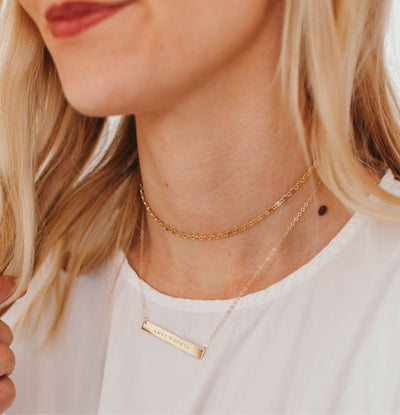 A close-up lifestyle shot of a blonde woman wearing a delicate gold bar engraved necklace on a fine chain, layered with a thicker gold chain.