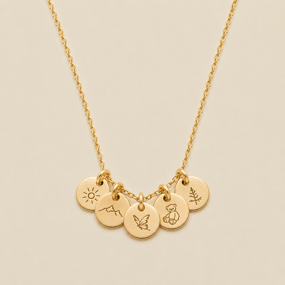 A gold chain necklace with five small round engraved charms featuring minimalist symbols of a sun, a mountain, butterfly, teddy bear, and pine tree, displayed on a light beige background.