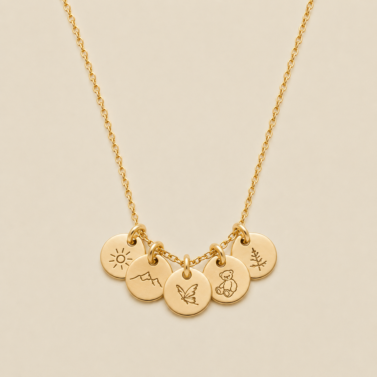 A gold chain necklace with five small round engraved charms featuring minimalist symbols of a sun, a mountain, butterfly, teddy bear, and pine tree, displayed on a light beige background.