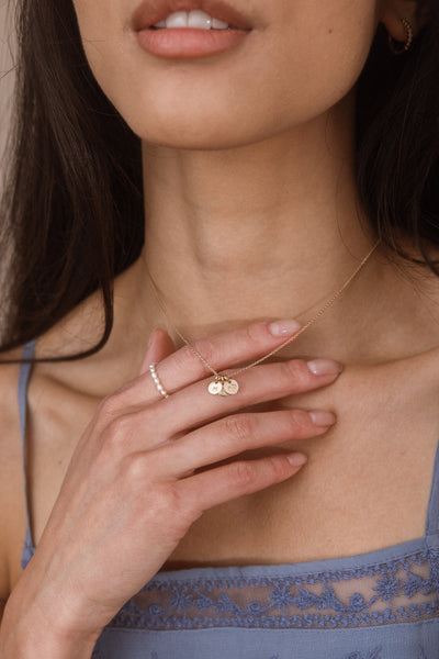 A close-up lifestyle shot of a model wearing a delicate gold necklace with three small round engraved charms and a gold ring on her finger.
