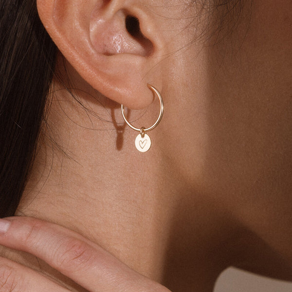 A close-up of a model's ear wearing a delicate gold hoop earring with a small round gold disc charm engraved with a heart.