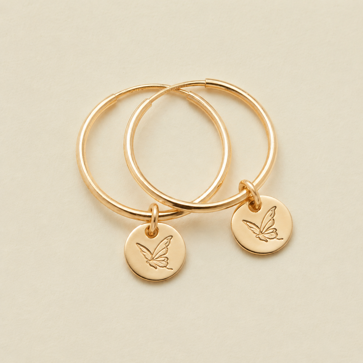 Two gold hoop earrings with small round engraved disc charms, each with a butterfly design, displayed on a light beige background.