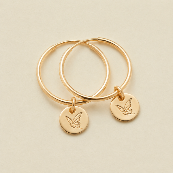Two gold hoop earrings with small round engraved disc charms, each with a butterfly design, displayed on a light beige background.