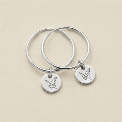 A pair of silver hoop earrings with small round engraved charms with a butterfly design, displayed on a light beige background.