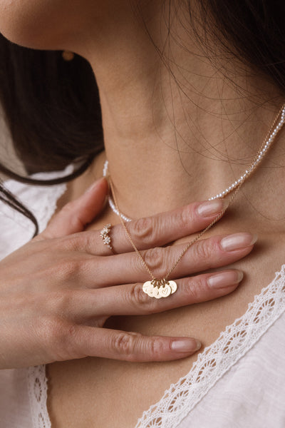 A close-up lifestyle shot of a woman wearing a delicate gold necklace with multiple engraved round disc pendants, layered over a pearl necklace, with a gold ring on her finger.