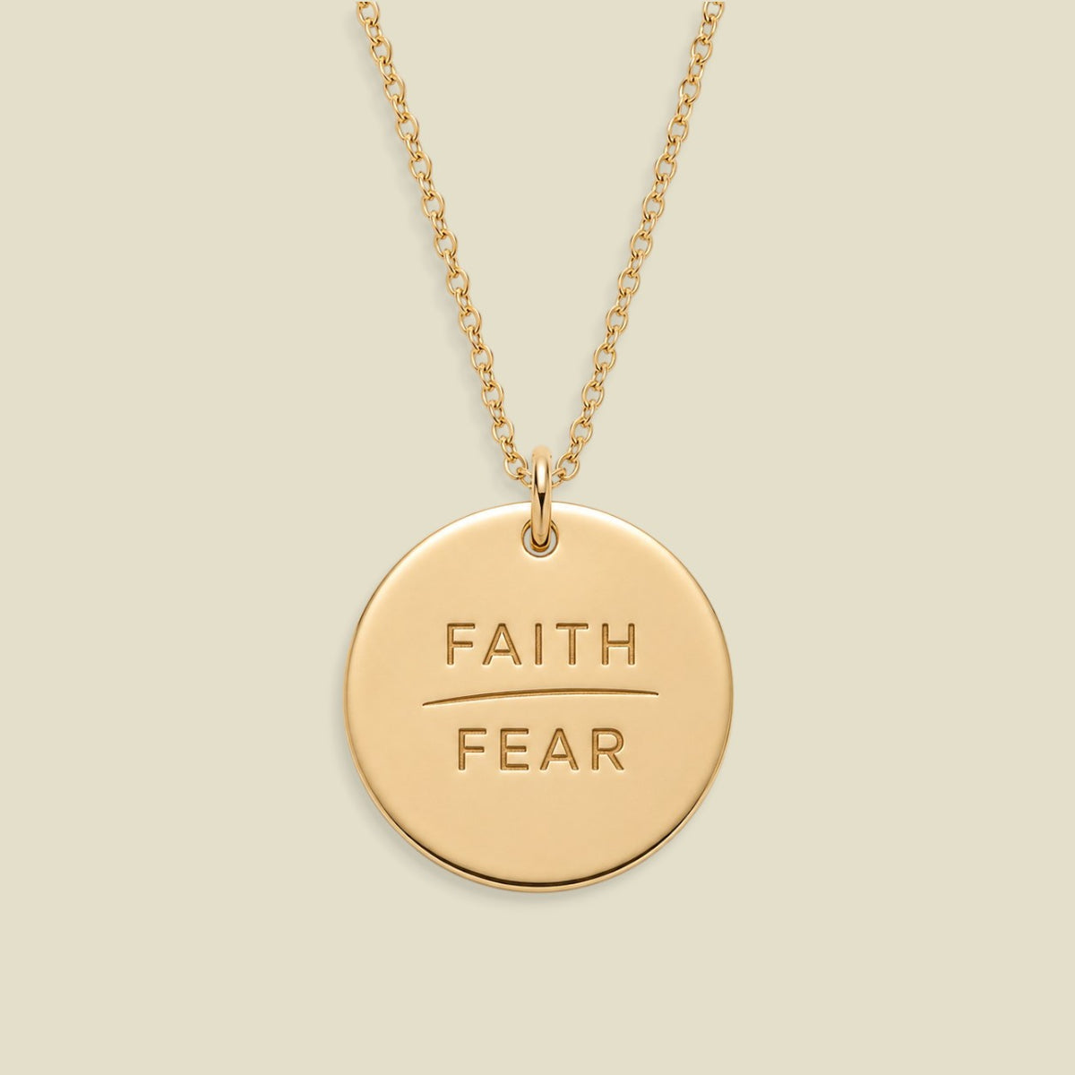 A gold disc pendant necklace engraved with the words 'FAITH' and 'FEAR' in capital letters, separated by a horizontal line, suspended from a delicate gold chain against a solid light beige background.