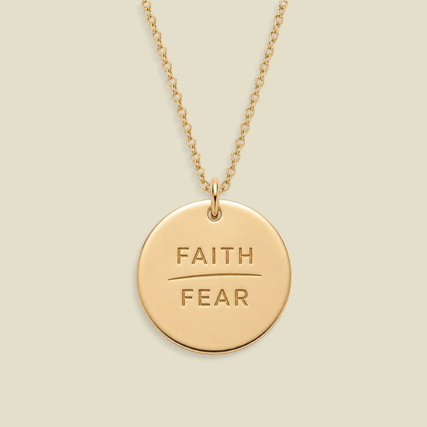 A gold disc pendant necklace engraved with the words 'FAITH' and 'FEAR' in capital letters, separated by a horizontal line, suspended from a delicate gold chain against a solid light beige background.