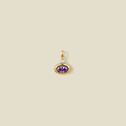 A gold oval pendant charm with a purple bezel-set oval gemstone and a clear accent stone on top, set against a solid beige background.