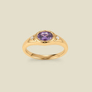 A gold signet ring with a purple center oval bezel-set gemstone, and two small clear accent stones on either side, set against a solid beige background.