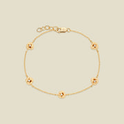 A delicate gold chain bracelet featuring five ornate, textured gold violet charms with small clear stones, arranged in a circle on a light beige background.