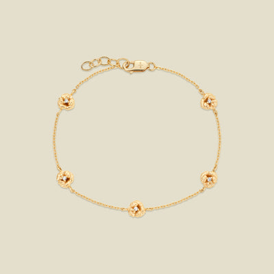 A delicate gold chain bracelet featuring five ornate, textured gold violet charms with small clear stones, arranged in a circle on a light beige background.