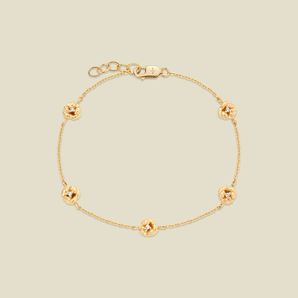 A delicate gold chain bracelet featuring five ornate, textured gold violet charms with small clear stones, arranged in a circle on a light beige background.