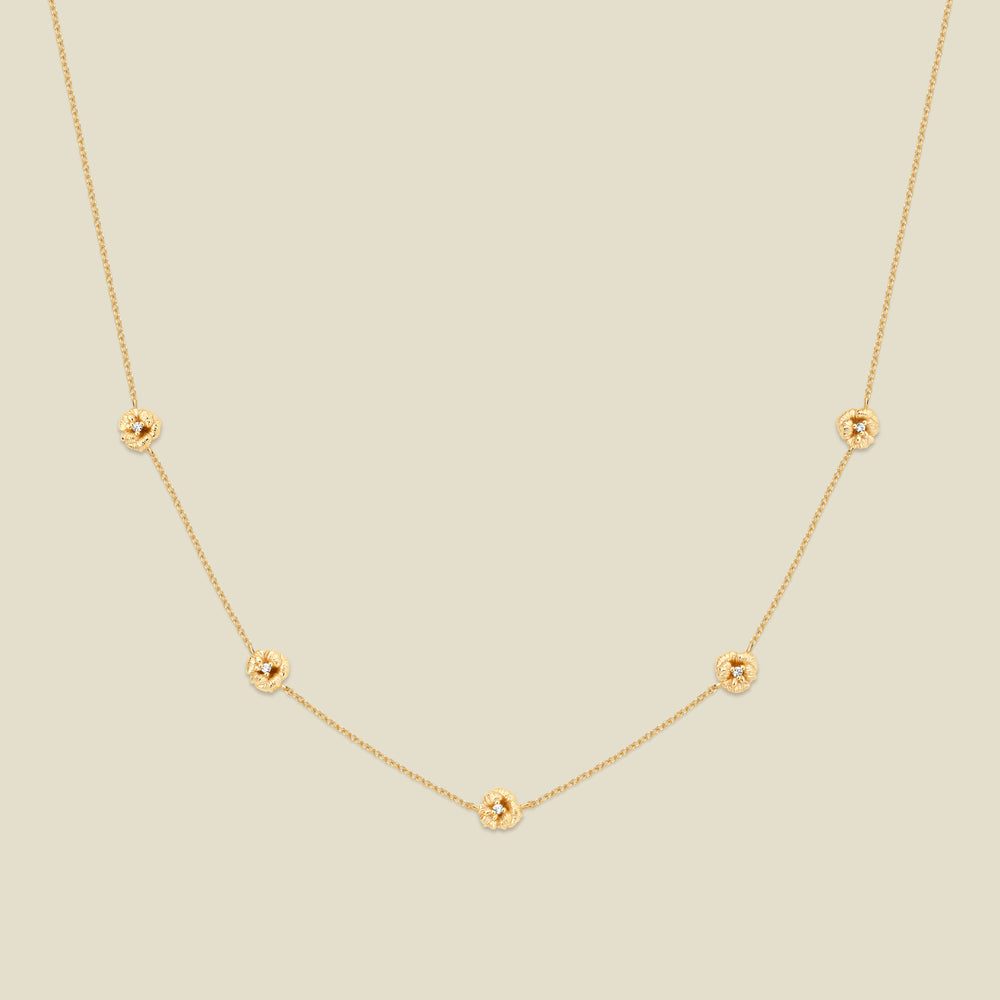 A delicate gold chain necklace featuring five small, evenly spaced textured violet flower motifs set with tiny clear stones, displayed on a solid light beige background.