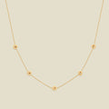 A delicate gold chain necklace featuring five small, evenly spaced textured violet flower motifs set with tiny clear stones, displayed on a solid light beige background.