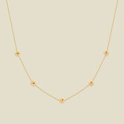 A delicate gold chain necklace featuring five small, evenly spaced textured violet flower motifs set with tiny clear stones, displayed on a solid light beige background.