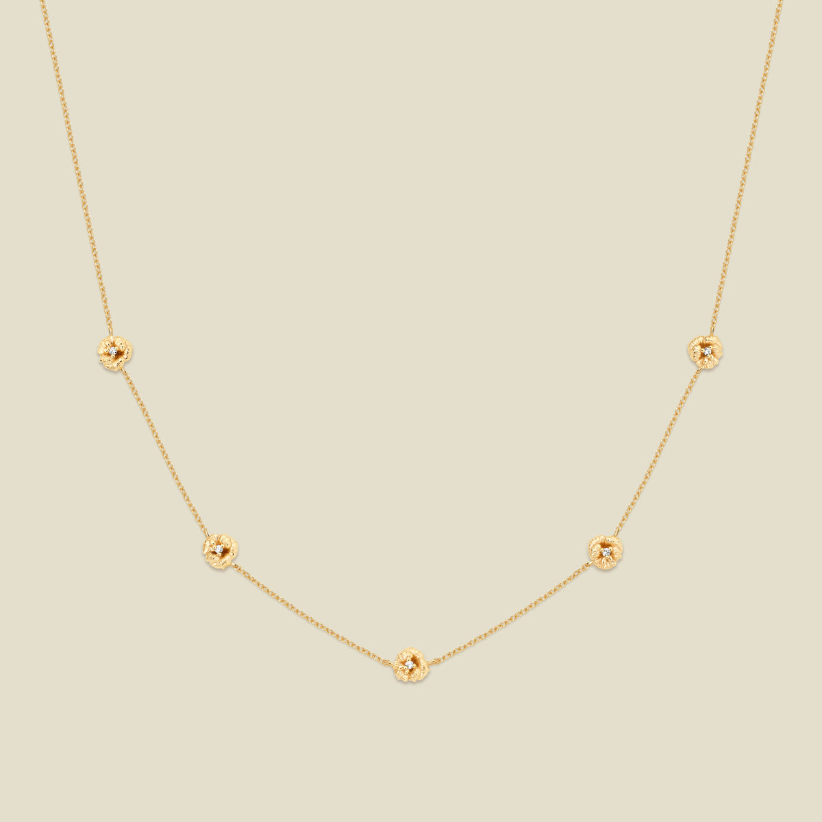 A delicate gold chain necklace featuring five small, evenly spaced textured violet flower motifs set with tiny clear stones, displayed on a solid light beige background.
