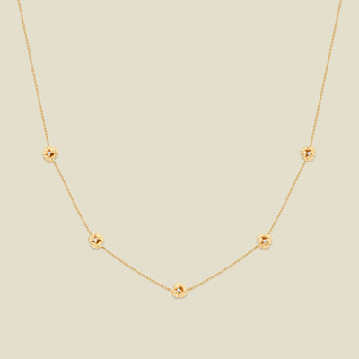 A delicate gold chain necklace featuring five small, evenly spaced textured violet flower motifs set with tiny clear stones, displayed on a solid light beige background.