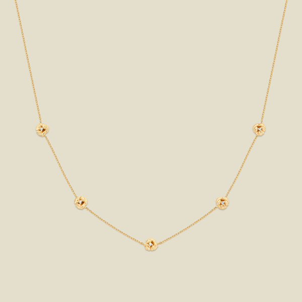 A delicate gold chain necklace featuring five small, evenly spaced textured violet flower motifs set with tiny clear stones, displayed on a solid light beige background.