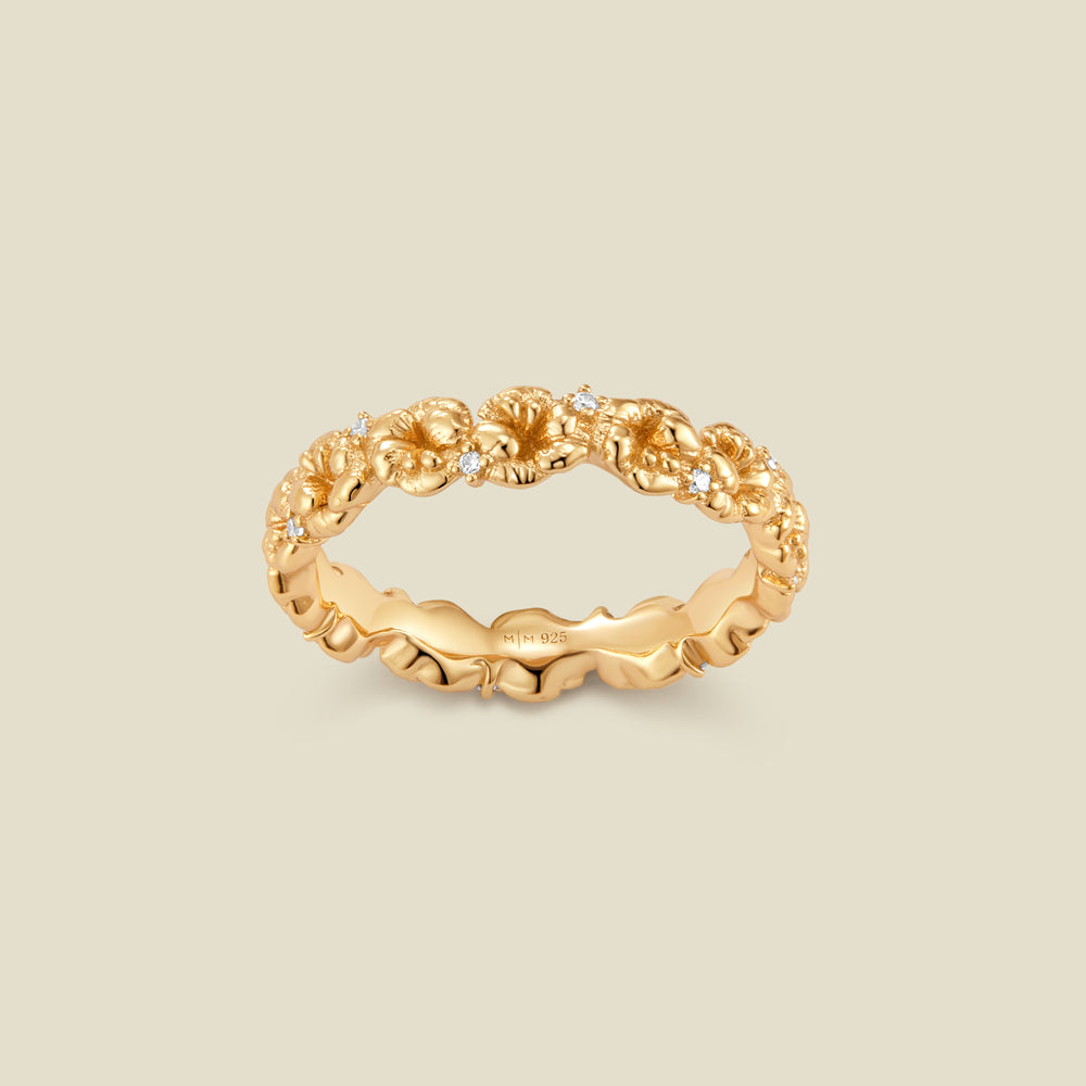 A gold floral-etched ring band with textured violet flowers and small clear gemstones, shown on a plain warm beige background. 