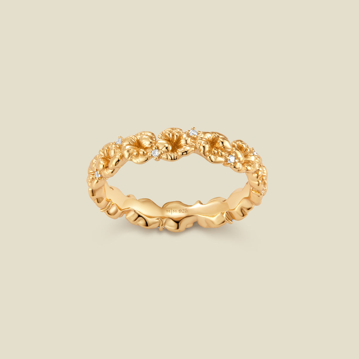 A gold floral-etched ring band with textured violet flowers and small clear gemstones, shown on a plain warm beige background. 