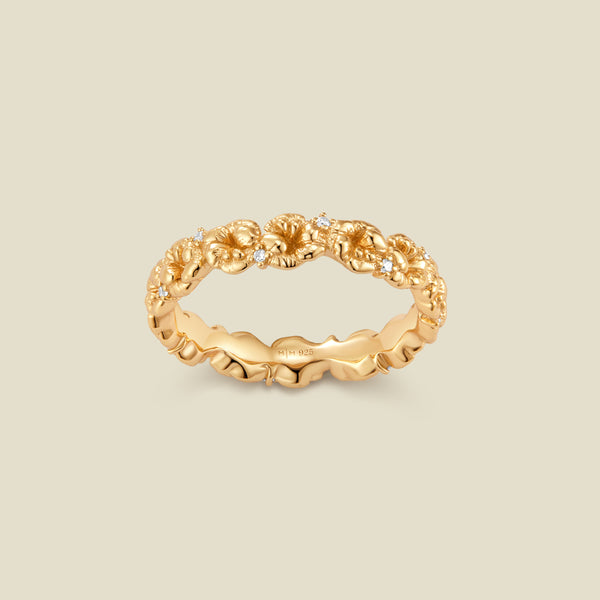 A gold floral-etched ring band with textured violet flowers and small clear gemstones, shown on a plain warm beige background. 