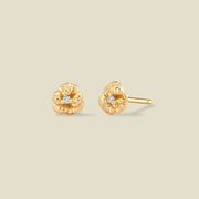 A pair of gold violet flower stud earrings, each featuring a small round clear gemstone at the center, displayed on a solid light beige background.