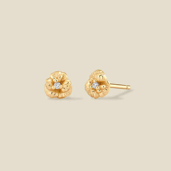 A pair of gold violet flower stud earrings, each featuring a small round clear gemstone at the center, displayed on a solid light beige background.