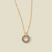 A gold chain necklace with a circular pendant featuring purple baguette-cut stones around a hollow center, displayed against a solid light beige background.
