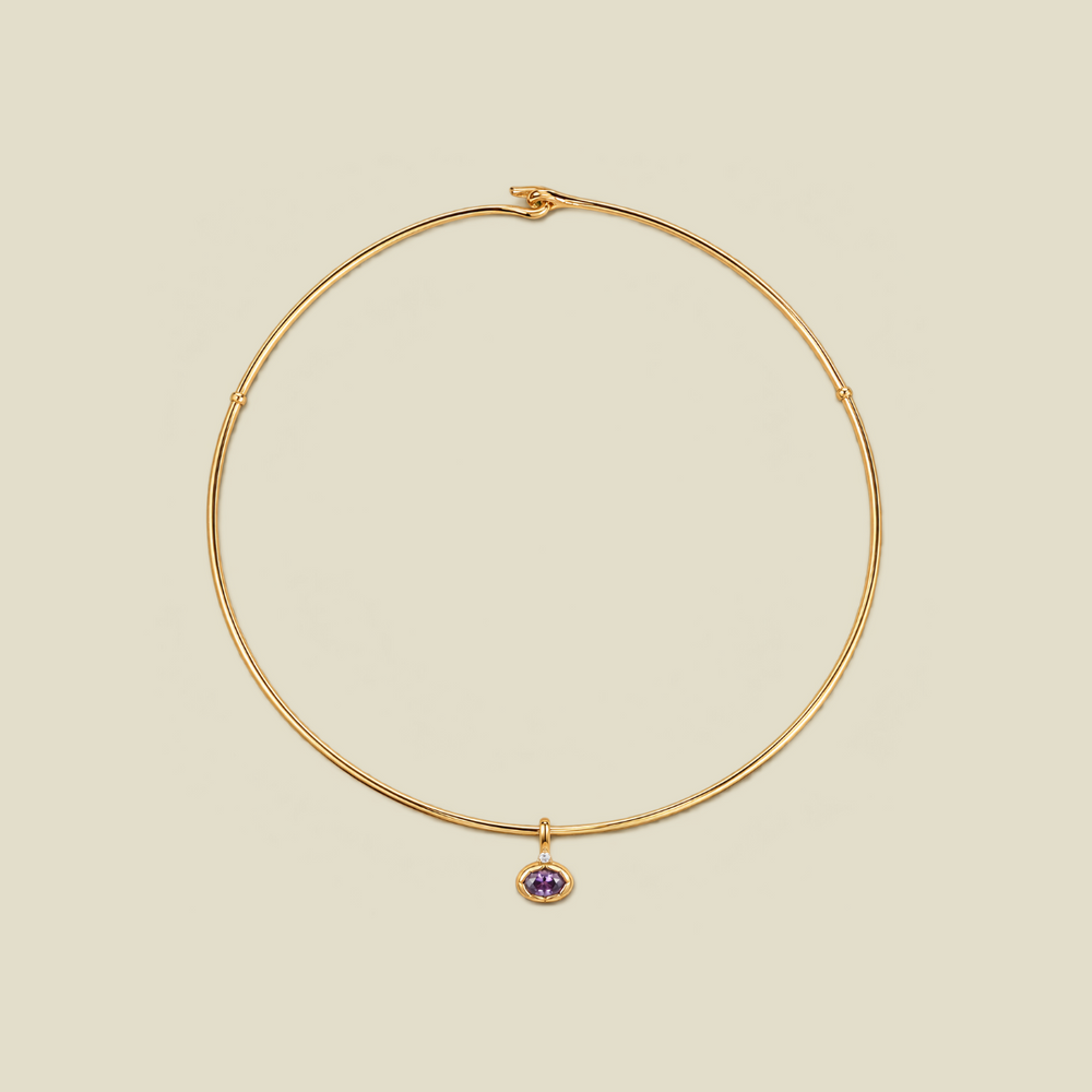 A thin gold wire necklace with a small purple oval gemstone charm, displayed on a solid beige background.