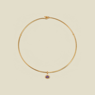 A thin gold wire necklace with a small purple oval gemstone charm, displayed on a solid beige background.