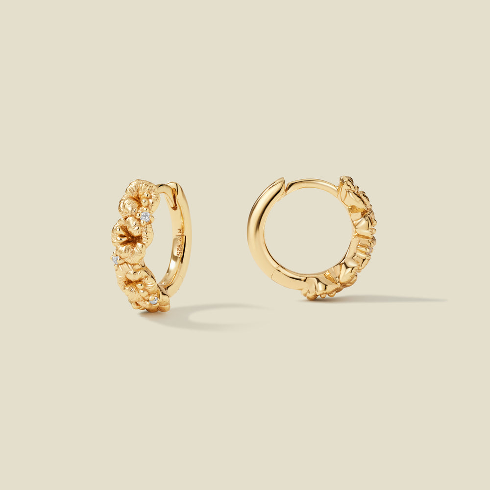 A pair of gold hoop earrings adorned with violet flowers stacked along the hoop, with scattered small clear accent stones, set against a solid beige background.