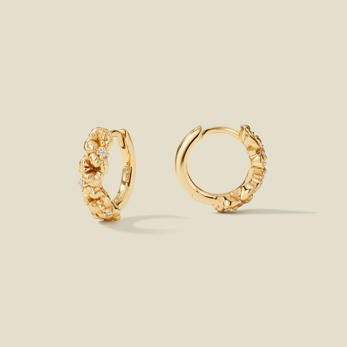 A pair of gold hoop earrings adorned with violet flowers stacked along the hoop, with scattered small clear accent stones, set against a solid beige background.