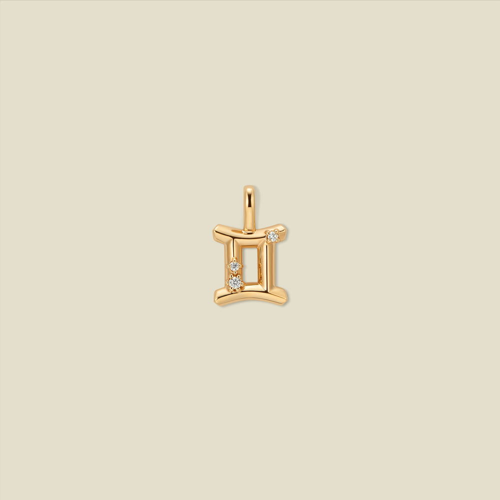 A gold pendant shaped like the Gemini zodiac symbol, featuring three small clear gemstones, centered on a solid light beige background.