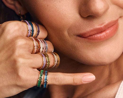 A close-up lifestyle shot of a model wearing a stack of colorful textured rings on her fingers, with her face partially visible in the background.