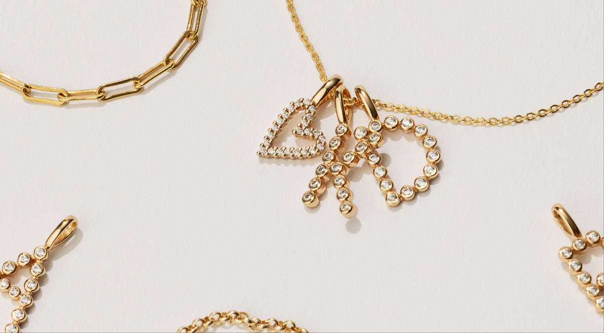 Gold-tone jewelry set featuring a central pendant necklace with a heart-shaped pavé ring and dangling letter charms, alongside matching chain necklaces and earrings on a light gray background.