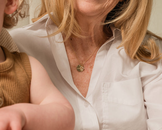 A close-up lifestyle shot of a woman wearing a delicate gold pendant necklace with a circular sun design, paired with a white button-down shirt. A child in a brown ribbed top is partially visible on the left.