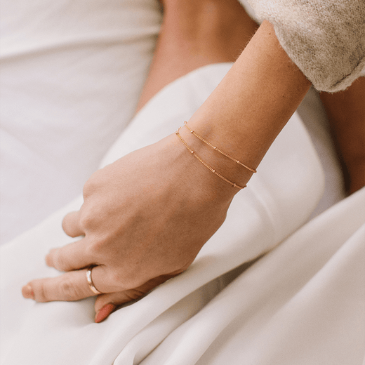 A person's wrist wearing two thin gold chain bracelets, each with stationary round polished beads.