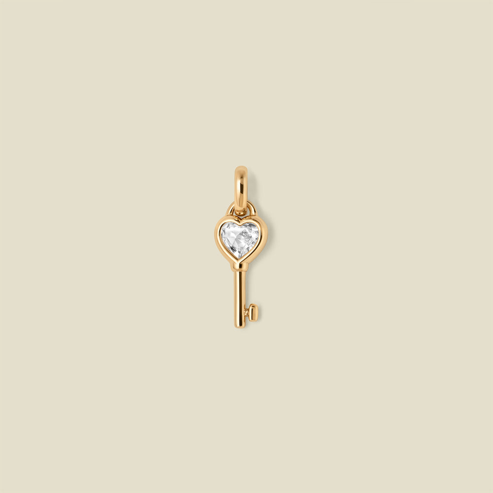 Gold key-shaped pendant with a heart shaped clear gemstone on a beige background